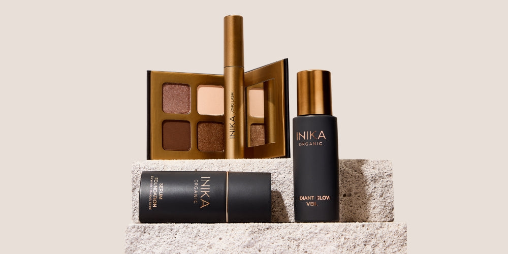 Luxury Australian makeup set with gold-bronze eyeshadow palette, mascara, radiant glow serum, and organic foundation on textured stone platform.