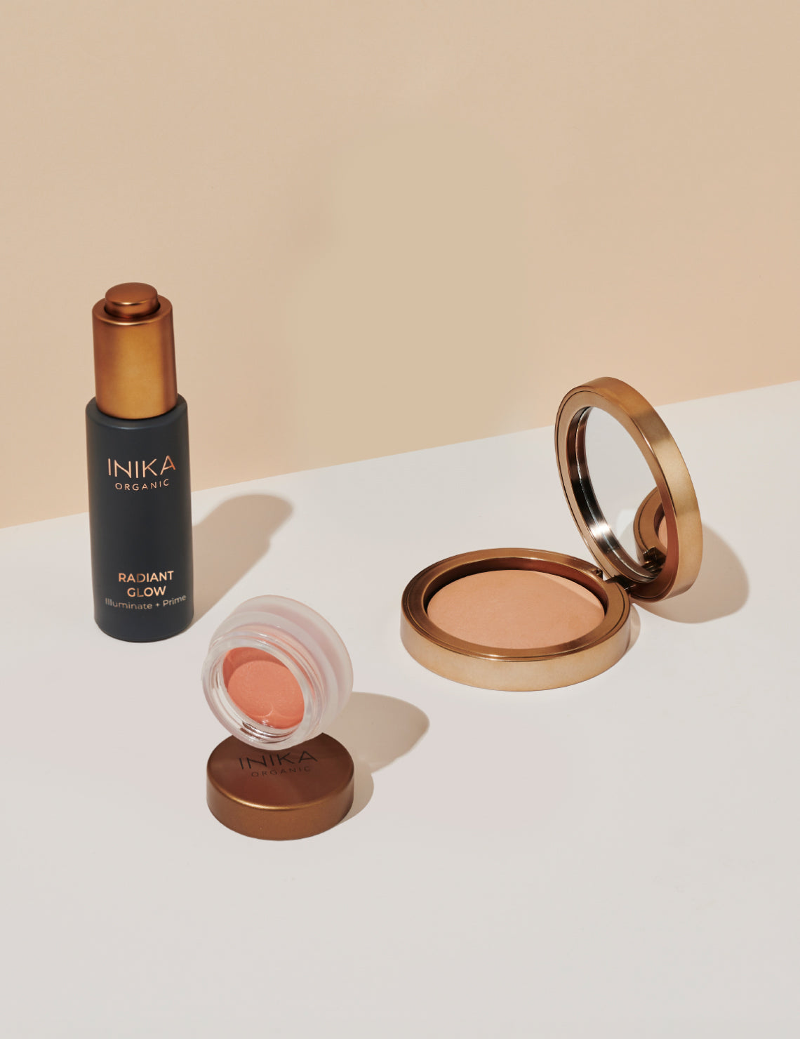 Inika Organic beauty products on a light background, including Radiant Glow serum, peach blush, and a bronzed compact powder.