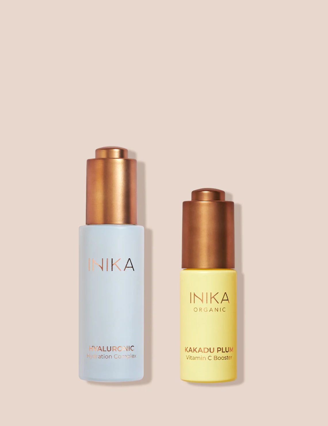 Inika Organics skincare: Hyaluronic Hydration Complex in blue bottle and Kakadu Plum Vitamin C Booster in yellow bottle, with copper caps, on peach background.